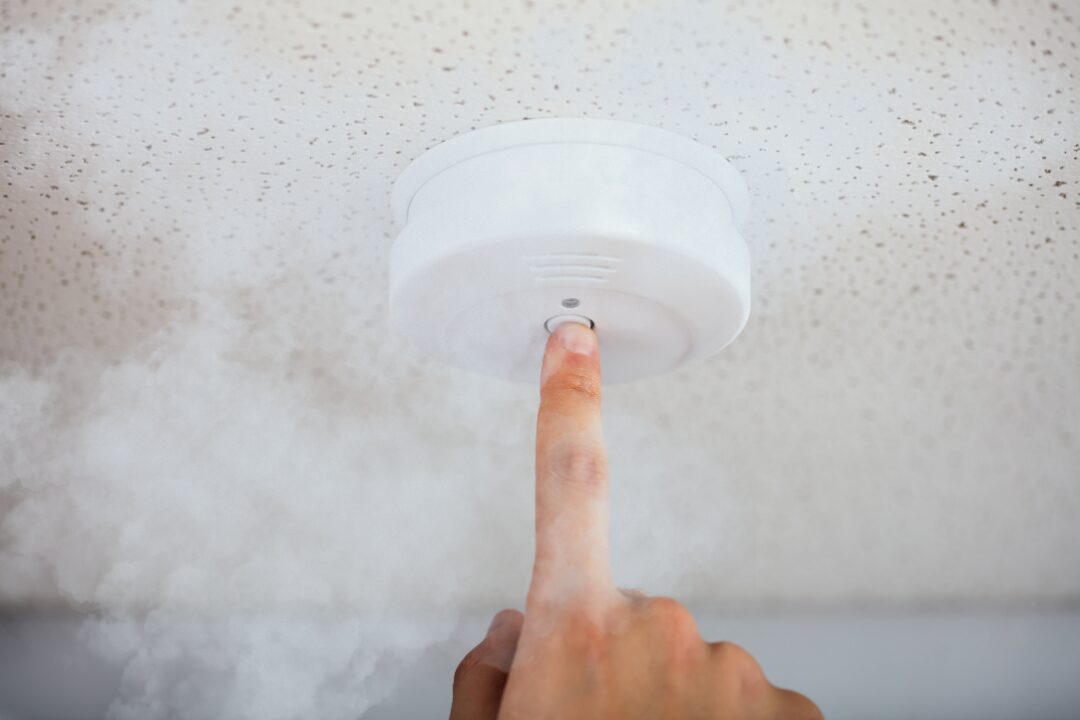 Finger pressing the button on a white smoke alarm on the ceiling, with smoke around it